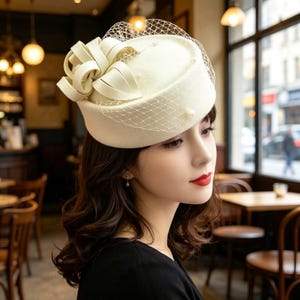 Pode incluir: Um chapéu pillbox de cor creme com uma base de feltro estruturada, um laço decorativo de fita na lateral e um delicado véu de rede. O chapéu é adornado com pequenas contas brancas redondas semelhantes a pérolas ao longo da coroa.