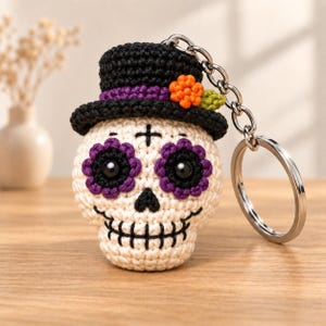 May include: A crocheted skull keychain featuring a cream-colored base, black stitched details, and purple eye sockets. The skull wears a black top hat adorned with a small orange flower and a green leaf, attached to a silver metal keyring.