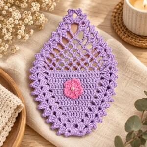 May include: A teardrop-shaped lavender crochet coaster featuring an intricate openwork lattice pattern on the top half and a solid stitch base on the bottom half. A small pink crocheted flower with a central white bead is attached to the centre.