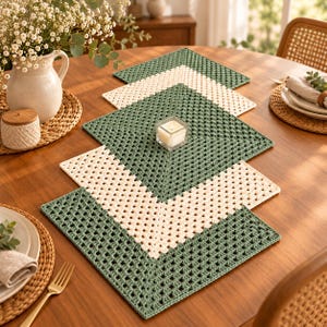 May include: A crochet table runner composed of alternating square motifs in sage green and cream yarn. The runner features a geometric granny square pattern arranged in a linear sequence, centered on a wooden table with a small square candle.