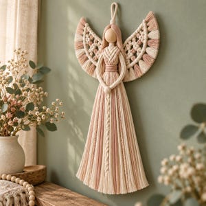 May include: A macrame wall hanging in the shape of an angel, crafted from cream and dusty pink cotton cord. The figure features a smooth wooden bead head, intricate knotted wings with fringed edges, and a long, flowing skirt with braided details.