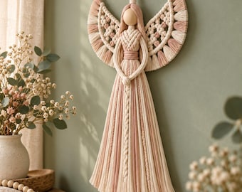 May include: A macrame wall hanging in the shape of an angel, crafted from cream and dusty pink cotton cord. The figure features a smooth wooden bead head, intricate knotted wings with fringed edges, and a long, flowing skirt with braided details.