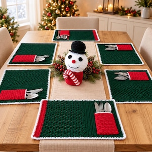 May include: A set of five rectangular dark green crochet placemats with white trim and a red pocket for silverware. A crocheted snowman figure with a black hat and red and white striped scarf sits in the center on a tan runner with pine decor.