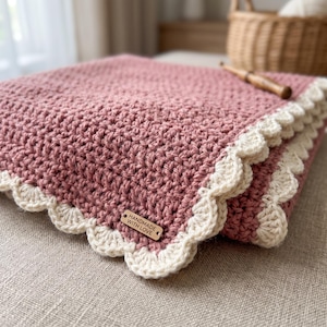 Puede incluir: Una manta de ganchillo color rosa empolvado con un patrón de punto texturizado y un borde festoneado de color crema. Una pequeña etiqueta rectangular de madera está fijada en la esquina, grabada con el texto "HANDMADE WITH LOVE" en letras negras.