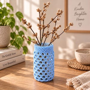Peut inclure: Un vase en verre transparent recouvert d'une housse en crochet bleu clair avec un motif ajouré répétitif. Le vase contient plusieurs branches ligneuses fines avec de petits bourgeons séchés brun clair pointant vers le haut.