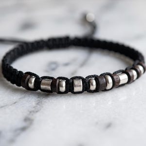 Puede incluir: Una pulsera de cordón negro con un patrón repetitivo de cuentas cilíndricas de madera marrón oscuro y cuentas de metal plateado pulido. La pulsera está construida con una técnica de nudos estilo macramé e incluye extremos ajustables.