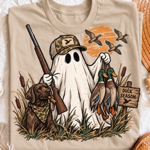 May include: A beige t-shirt featuring a graphic of a ghost wearing a camouflage baseball cap and holding a shotgun. A brown dog with a camouflage bandana sits nearby. Two ducks hang from the ghost's hand, with more ducks flying above a wooden sign reading DUCK SEASON.