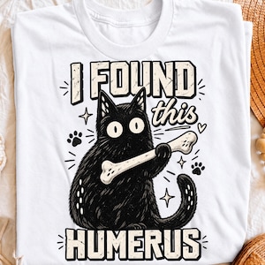 May include: A white t-shirt featuring a black cat illustration holding a bone. The text reads I FOUND this HUMERUS in bold, distressed lettering. The design includes small star, heart, and paw print accents surrounding the cat, which has wide eyes.