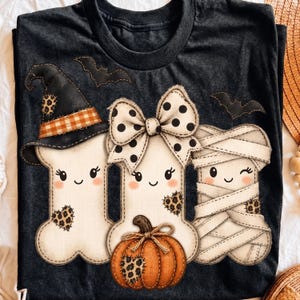 May include: A dark gray t-shirt featuring a graphic of three bone-shaped characters. One wears a black witch hat, one has a large white and black polka dot bow, and one is wrapped like a mummy. An orange pumpkin sits below with leopard print hearts.