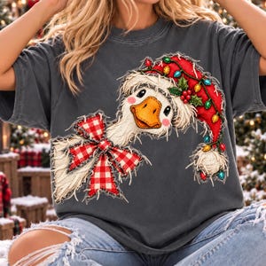 May include: A charcoal grey t-shirt featuring a graphic of a white goose wearing a red Santa hat decorated with colourful Christmas lights. The goose has a red and white chequered bow tied around its neck, with a textured, frayed appearance.
