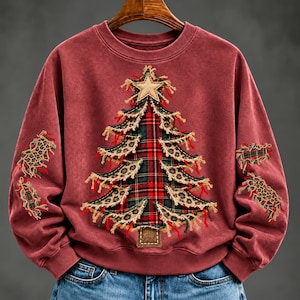 Puede incluir: Una sudadera de cuello redondo color granate con un gran aplique de árbol de Navidad central hecho de tela de cuadros rojos y verdes, con bordes deshilachados de estampado de leopardo y una estrella beige en la punta. Las mangas tienen parches a juego.