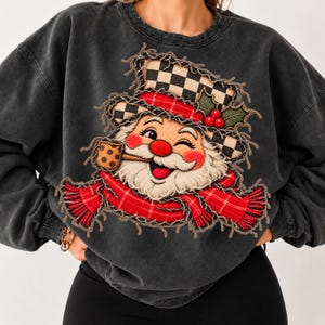 Puede incluir: Una sudadera gris carbón de cuello redondo con una gran aplicación de bordes deshilachados de un Santa Claus guiñando un ojo. Santa lleva un gorro de cuadros blancos y negros con bayas de acebo rojas y una bufanda de cuadros rojos.