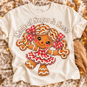 May include: An off-white t-shirt featuring a graphic of a gingerbread person with brown skin, wearing a red and white checkered dress and matching hair bows. The text "Made of Sugar & Sass!" is printed in a brown, arched font above the character.