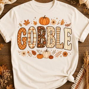 Puede incluir: Una camiseta de manga corta color crema con la palabra GOBBLE en letras grandes y estampadas. Cada letra tiene un diseño único que incluye lunares, cuadros, flores y rayas. El gráfico incluye calabazas naranjas y hojas de otoño.