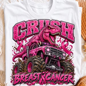 May include: A white t-shirt featuring a graphic of a pink Tyrannosaurus Rex standing on a pink monster truck. The text CRUSH BREAST CANCER is displayed in bold pink letters, with pink awareness ribbons integrated into the design and mud splatters.