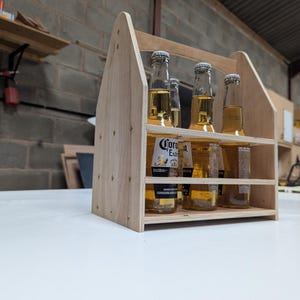May include: A light-coloured wooden beer caddy with a handle and open sides, holding four glass bottles of Corona Extra beer. The carrier is constructed from plywood panels joined with visible screws, designed to transport standard beverage bottles.