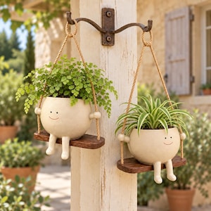 May include: Two off-white ceramic planters with simple smiling faces, dangling legs, and arms, each resting on a small dark brown wooden swing. The planters are suspended by tan ropes from a dark metal wall-mounted hook attached to a wooden post.