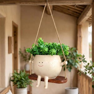 May include: A round, cream-colored ceramic planter with a simple smiling face, featuring two small arms and legs, sitting on a small wooden swing suspended by two braided ropes, containing a vibrant green succulent plant with dense, rounded leaves.