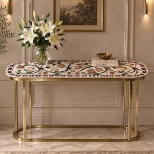 May include: An oval console table with a gold-toned metal frame and a white stone top featuring an inlaid floral pattern in shades of red, green, and brown. A glass vase with white lilies and two stacked books sit on the table surface.