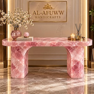 May include: A rectangular console table crafted from polished pink rose quartz with a smooth surface and two arched legs. The table is topped with a glass vase of white flowers, a book titled The Kinfolk, and a gold tray with a candle and diffuser.