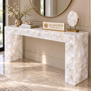 Può includere: Un tavolo console rettangolare costruito con pezzi di pietra di quarzo bianco traslucido. Sulla superficie si trovano un cartello rettangolare con il testo ALAFUWW HANDICRAFTS, una scultura circolare in quarzo su supporto dorato, una ciotola e libri.