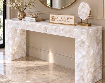 May include: A rectangular console table constructed from translucent white quartz stone pieces. On the surface sits a rectangular sign with the text ALAFUWW HANDICRAFTS, a circular quartz sculpture on a gold stand, a small bowl, and books.