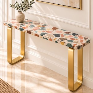 Pode incluir: Uma mesa consola longa e estreita com um tampo retangular apresentando um padrão de mosaico multicolorido de fragmentos de pedra em tons de rosa, verde, castanho e preto. A mesa é suportada por duas pernas em forma de U com acabamento dourado.