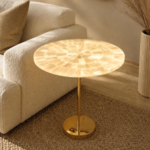 May include: A round side table featuring a translucent, cream-colored stone top with a radial mosaic pattern. The table is supported by a single, slender, polished gold-tone metal pedestal base that connects to a circular, flat gold-tone metal floor plate.