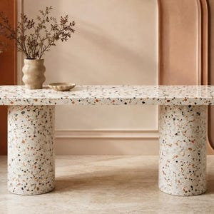 May include: A rectangular console table with a terrazzo finish featuring a white base with flecks of orange, brown, and black. The table has a smooth top with rounded corners and is supported by two thick, cylindrical legs made of the same material.