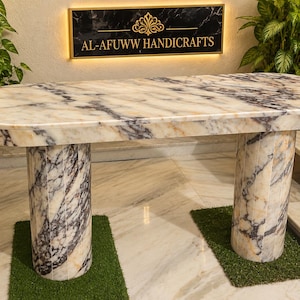 May include: An oval marble table featuring white, gray, and gold veining throughout the surface and two cylindrical pedestal bases. A black sign with gold lettering reads AL-AFUWW HANDICRAFTS with a decorative gold emblem above the text.
