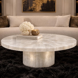 May include: A round coffee table featuring a white, translucent, crystalline stone surface with a radial pattern and a matching cylindrical pedestal base. A white book titled The Kinfolk, a white floral arrangement, and a gold bowl sit on top.