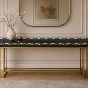 May include: A rectangular console table featuring a black top with a white floral inlay border and a gold-toned metal frame. A cream-colored ceramic vase containing white flowering branches and a small gold sphere sit on the table surface.