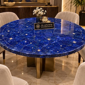 Può includere: Un tavolo da pranzo rotondo con un piano in pietra lapislazzuli blu lucido con inserti a forma di stella dorata. Il tavolo presenta una base centrale in metallo color oro e un cartello nero con la scritta AL-AFUWW HANDICRAFTS.