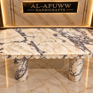 May include: An oval dining table made of white and dark purple veined marble with a scalloped edge. The table is supported by two thick cylindrical marble legs. A sign on the wall behind the table reads AL-AFUWW HANDICRAFTS.