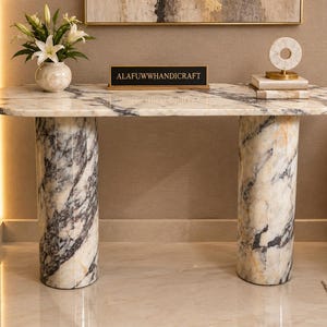 Op de afbeelding: Een ovale consoletafel van wit en grijs geaderd marmer, voorzien van twee dikke, cilindrische, gecanneleerde poten. Een zwart rechthoekig bord met de gouden tekst ALAFUWWHANDICRAFT staat op het oppervlak, naast een witte vaas en boeken.