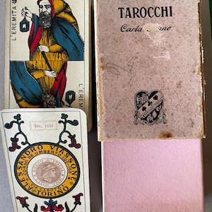 Puede incluir: Un mazo de cartas de juego vintage Tarocchi que incluye la carta L'Eremita con una figura barbuda sosteniendo una linterna. La caja está etiquetada como Tarocchi y A. Viassone-Torino. Una carta separada muestra un emblema circular con el texto Alessandro Viassone Torino.