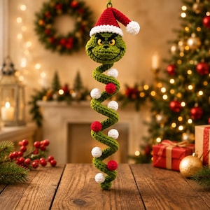 May include: A crocheted green character ornament with a frowning expression, wearing a red and white Santa hat. The body is a long, green spiral shape decorated with small, alternating red and white crocheted spheres attached along the length.