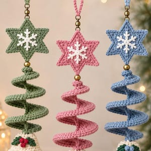 May include: Three hanging crochet ornaments in sage green, dusty pink, and light blue. Each features a six-pointed star with a white snowflake, a spiral tree body, and a cream-colored bell base with small green leaves and berries, finished with a brass bell.