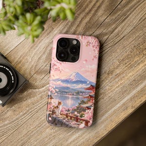 以下が含まれることがあります： スマートフォンケース。湖、富士山、桜に囲まれた五重塔を見下ろす石垣の上に座る猫のイラストがプリントされています。デザインには、ソフトなピンク、白、茶色の色調が使われています。
