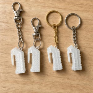 May include: Four keychains featuring translucent white, textured, pistol-grip shaped charms. Two keychains have silver-toned lobster claw clasps, one has a gold-toned split ring, and one has a silver-toned split ring, all attached by metal chains.