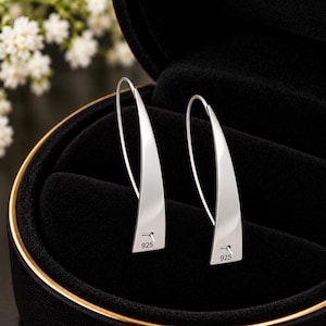 Può includere: Un paio di orecchini pendenti in argento dal design curvo e allungato. Ogni orecchino presenta una superficie liscia e lucida ed è stampigliato con il testo 925 nella parte inferiore, indicando una composizione in argento sterling.