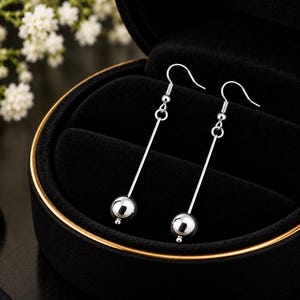 May include: A pair of silver-toned drop earrings featuring a long, thin vertical bar suspended from a hook wire, with a single polished spherical bead attached to the bottom end of each bar, showcasing a clean, reflective metallic finish.