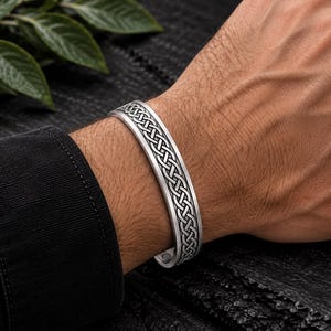 May include: A silver-toned metal cuff bracelet featuring an intricate, repeating Celtic knot pattern engraved along the centre. The bracelet has a polished finish with darkened recessed areas to highlight the detailed woven design.