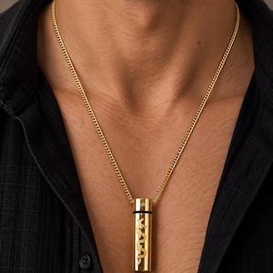 May include: A gold-toned cylindrical pendant necklace featuring a cut-out snake design and a small embedded clear stone. The pendant hangs from a matching gold-toned curb chain, resting against a black collared shirt. The metal has a polished finish.