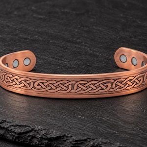 Puede incluir: Una pulsera de cobre tipo brazalete con un patrón de nudo celta en relieve a lo largo de la banda exterior. El interior de la pulsera muestra dos imanes circulares incrustados en cada extremo, con un acabado metálico y detalles trenzados.