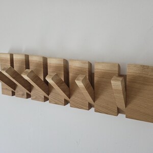 Könnte beinhalten: Eine Wandgarderobe aus hellem Holz mit natürlicher Maserung. Die Garderobe besteht aus mehreren rechteckigen Paneelen, von denen einige als schräge Haken ausgeklappt werden können, während andere flach an der Basis anliegen.