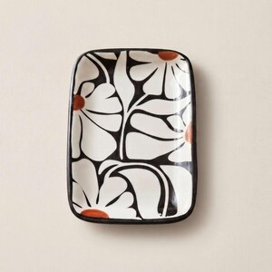 Può includere: Un vassoio rettangolare in ceramica con angoli arrotondati caratterizzato da un audace motivo floreale bianco e nero. Il design è composto da grandi petali di fiori bianchi stilizzati delineati in nero, con piccoli centri color terracotta.