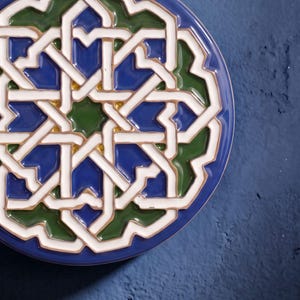 Könnte beinhalten: Eine kreisförmige Keramikfliese mit einem komplexen geometrischen Sternmuster in Weiß, Dunkelblau und Waldgrün. Das Design besteht aus ineinandergreifenden Linien und Formen mit einer glänzenden Oberfläche, die ein symmetrisches Motiv bilden.