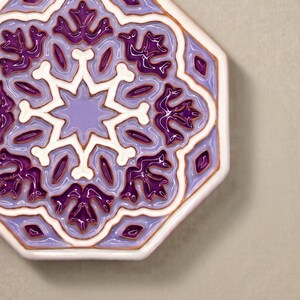 Puede incluir: Un azulejo de cerámica octogonal con un patrón en relieve brillante en tonos morado oscuro, lavanda claro y blanco. El diseño consiste en una estrella central rodeada de motivos florales simétricos y bordes geométricos definidos.