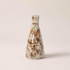 Può includere: Un vaso in ceramica dalla forma conica affusolata e collo stretto. La superficie presenta uno smalto lucido color bianco sporco ricoperto da un fitto motivo maculato di marrone e marrone chiaro che varia per dimensioni e intensità.